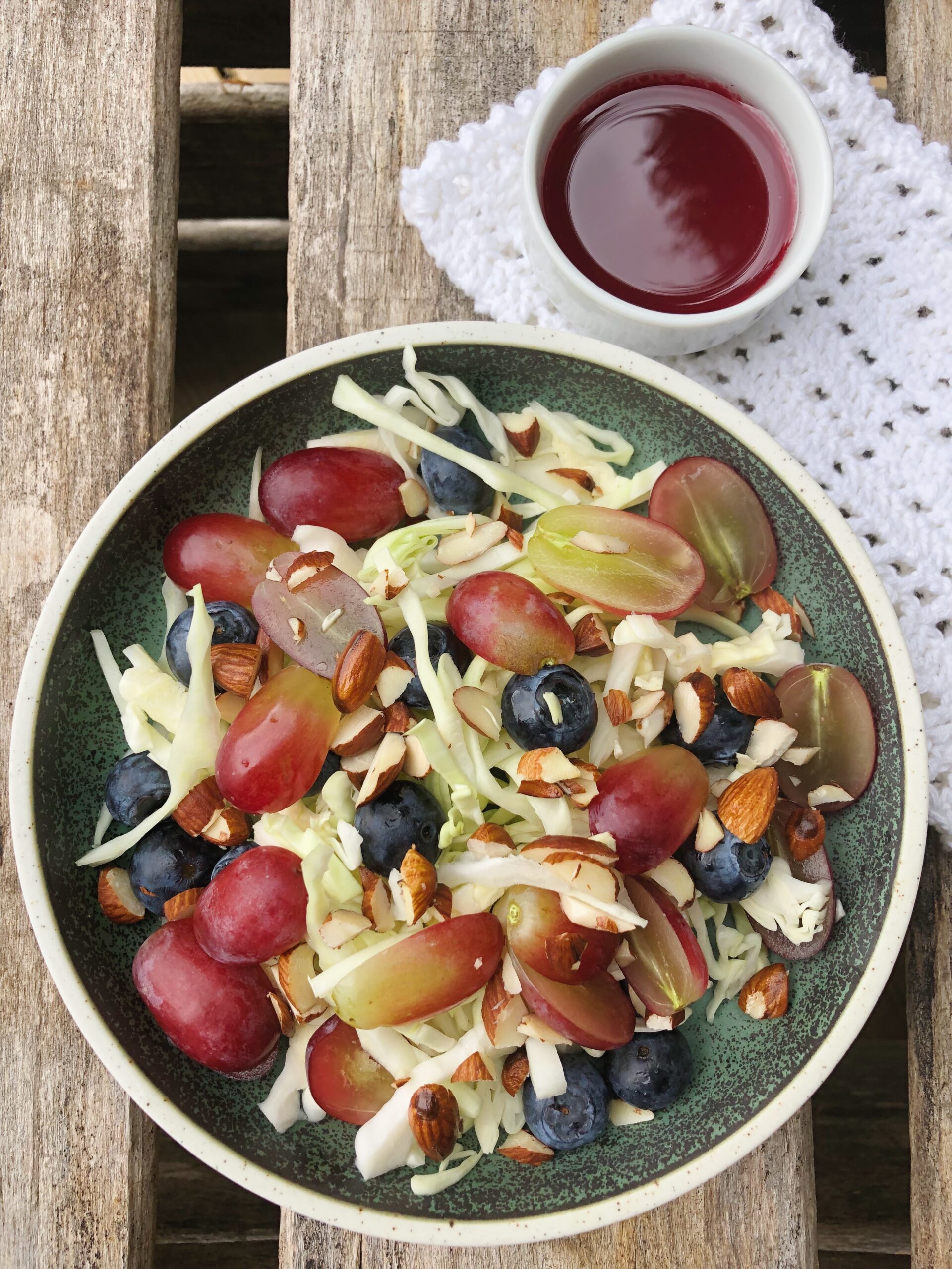 Spidskålsalat med rabarbersirup i en grøn tallerken som står på en trækasse, hvor der står en lille skål med rabarbersirup ved siden af