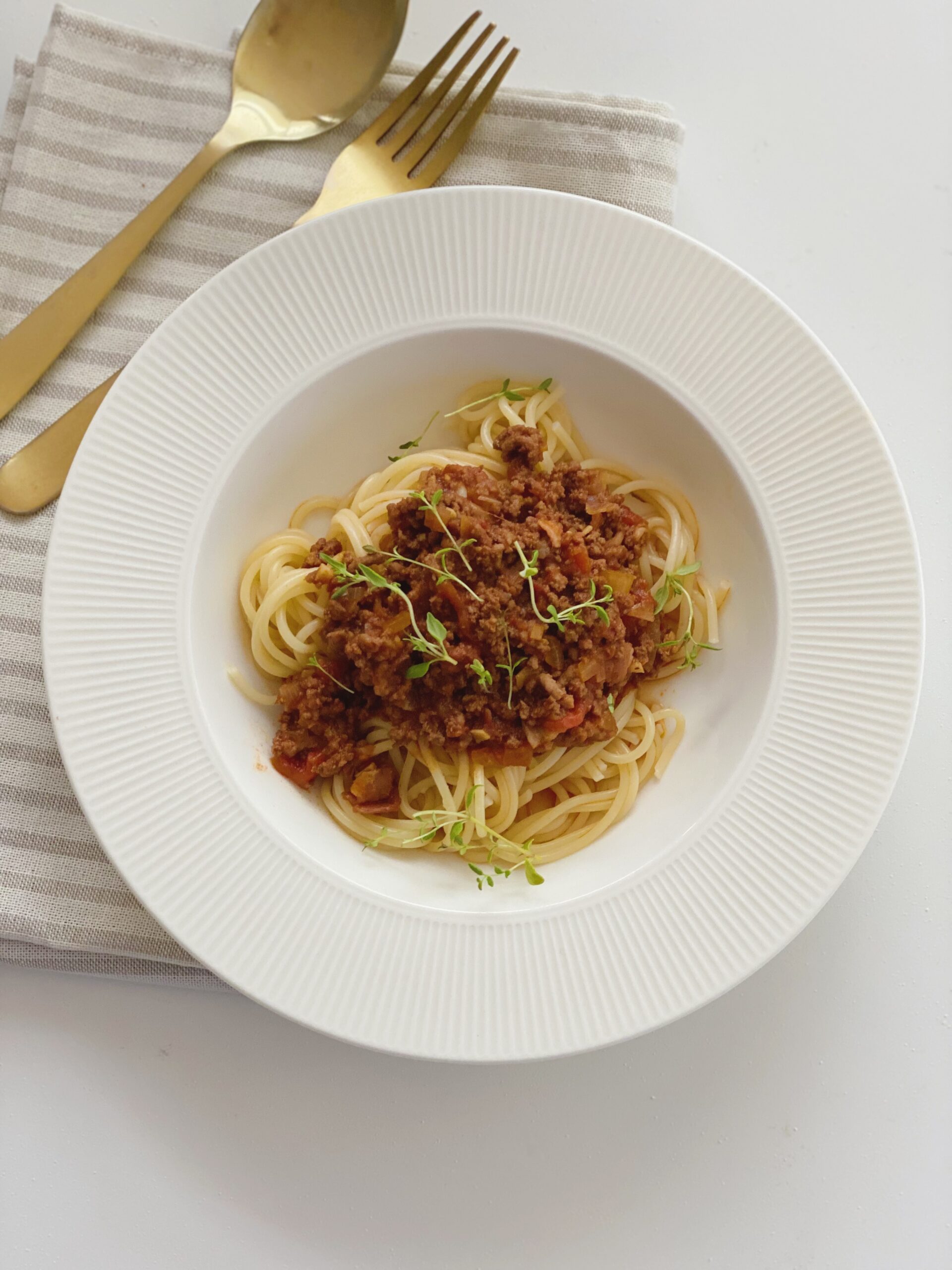 Spaghetti bolognese set oppe fra, hvor hele tallerkenen er synlig og der ligger en ske og en gaffel ved siden af oven på en stribet serviet